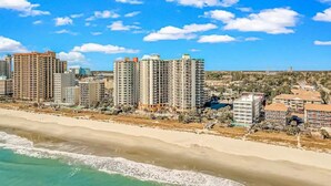 A coastal cityscape with high-rise buildings along the beach, clear skies, and a sandy shoreline.
