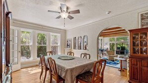 Une salle à manger avec une table dressée pour quatre personnes, des chaises en bois, un ventilateur de plafond et de grandes fenêtres.