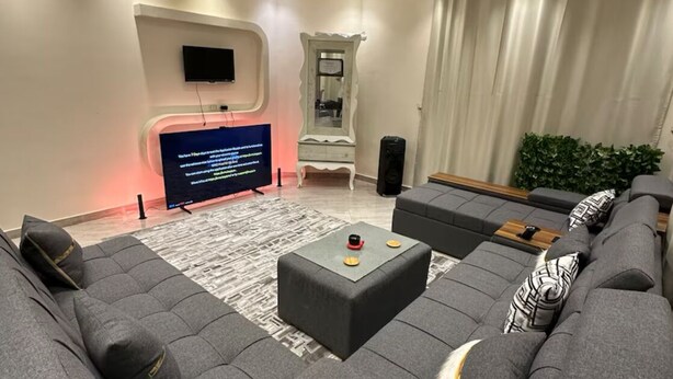 A modern living room with a sectional sofa, a flat-screen TV, a decorative mirror, and a large window with curtains.