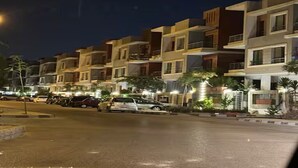 A row of modern residential buildings with balconies, a street in the foreground, and parked cars.