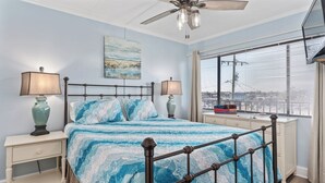 A bedroom with a bed, two bedside tables, a ceiling fan, a window with blinds, and a city view. - Beachfront Boardwalk Escape (Carolina Beach)