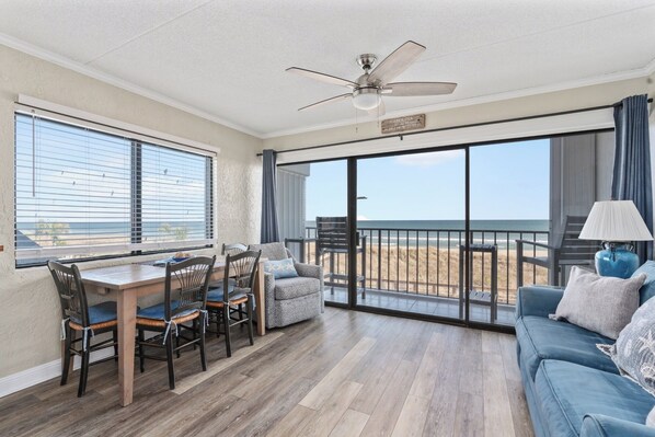 A spacious balcony with a view of the beach, a dining area with chairs and a table, and a ceiling fan.