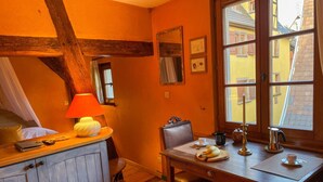 A cozy room with a wooden table, a chair, a lamp, and a window with a view of buildings outside. - La Maison Bleue - Historic house in the heart of Riquewihr. "Le Renard" 2-guest apartment (Riquewihr)
