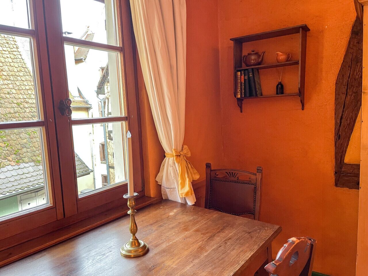 Une pièce avec une table en bois, une chaise, une fenêtre avec des rideaux et une étagère contenant des livres et une théière.