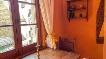 Une pièce avec une table en bois, une chaise, une fenêtre avec des rideaux et une étagère contenant des livres et une théière.