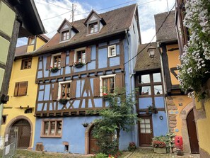 A traditional half-timbered house with a steeply pitched roof, dormer windows, and a small garden area with potted plants and flowers. - La Maison Bleue - Historic house in the heart of Riquewihr. "Le Renard" 2 guest flat (Riquewihr)