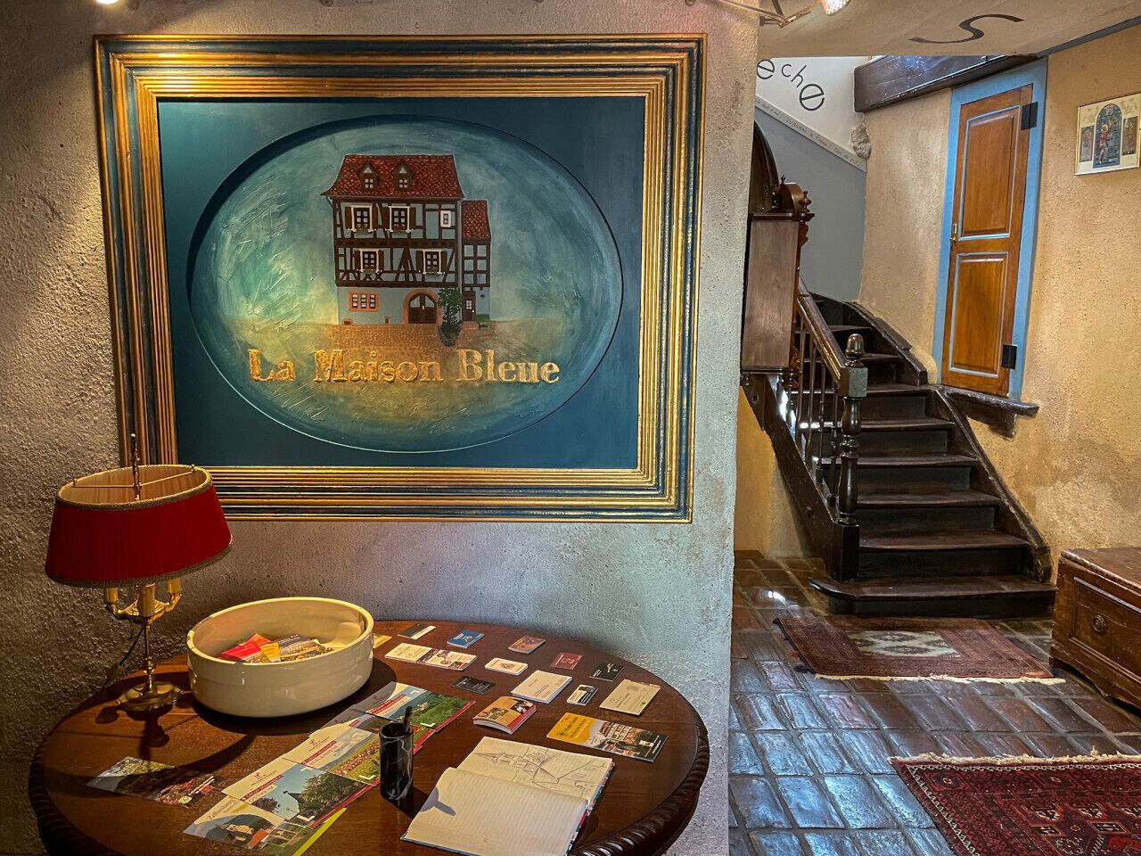 Un hall d’hôtel avec un tableau encadré représentant une maison, un escalier et une table ronde sur laquelle sont disposées des brochures.