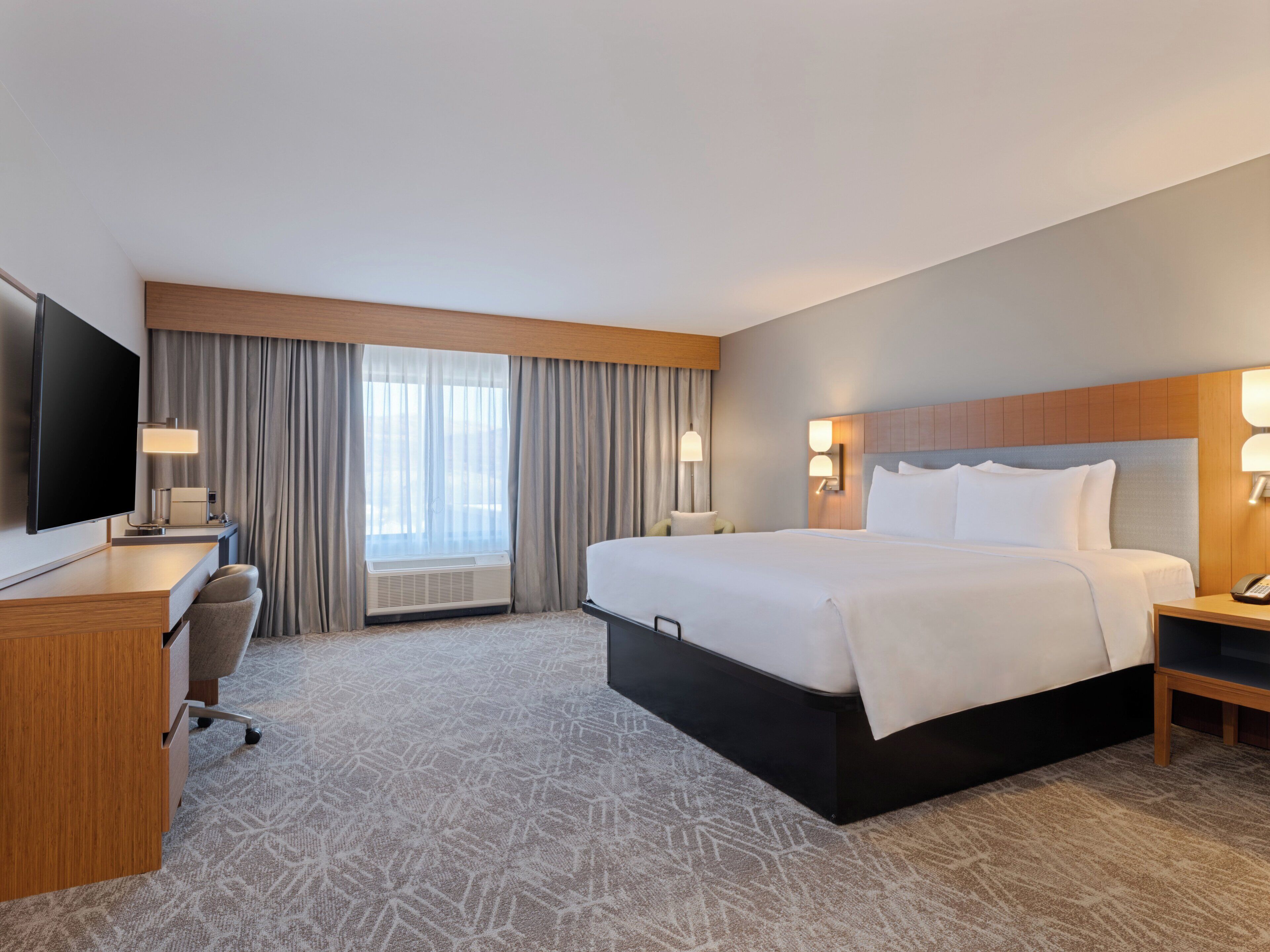 A hotel room with a large bed, a desk with a flat-screen TV, a chair, a bedside table with a lamp, and a window with curtains.