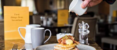 A breakfast setting with pancakes topped with bacon and butter, a coffee cup, and a sugar dispenser on a marble table.