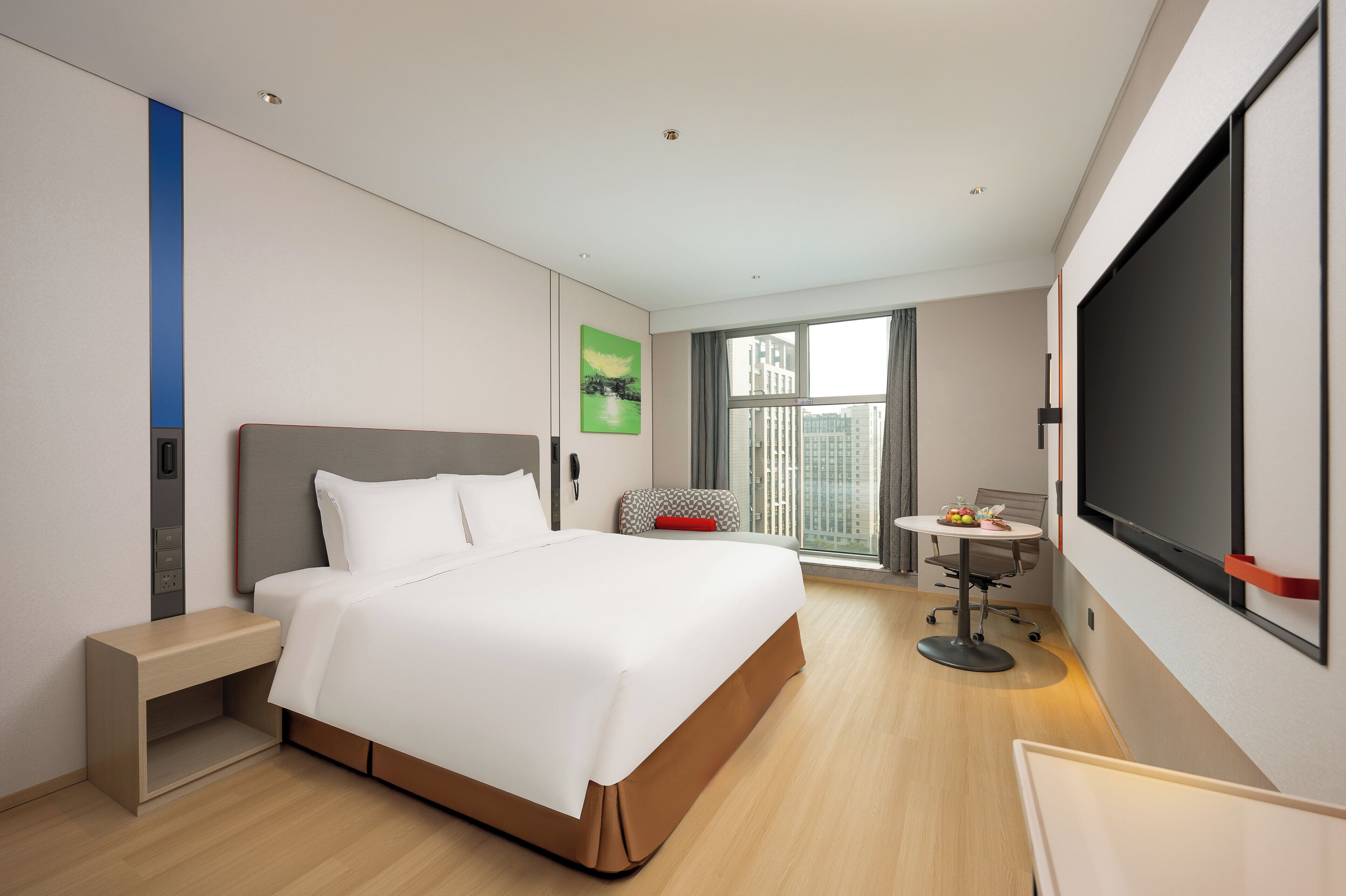 A modern hotel room with a bed, bedside table, a desk with a chair, a large window, and a flat-screen TV.
