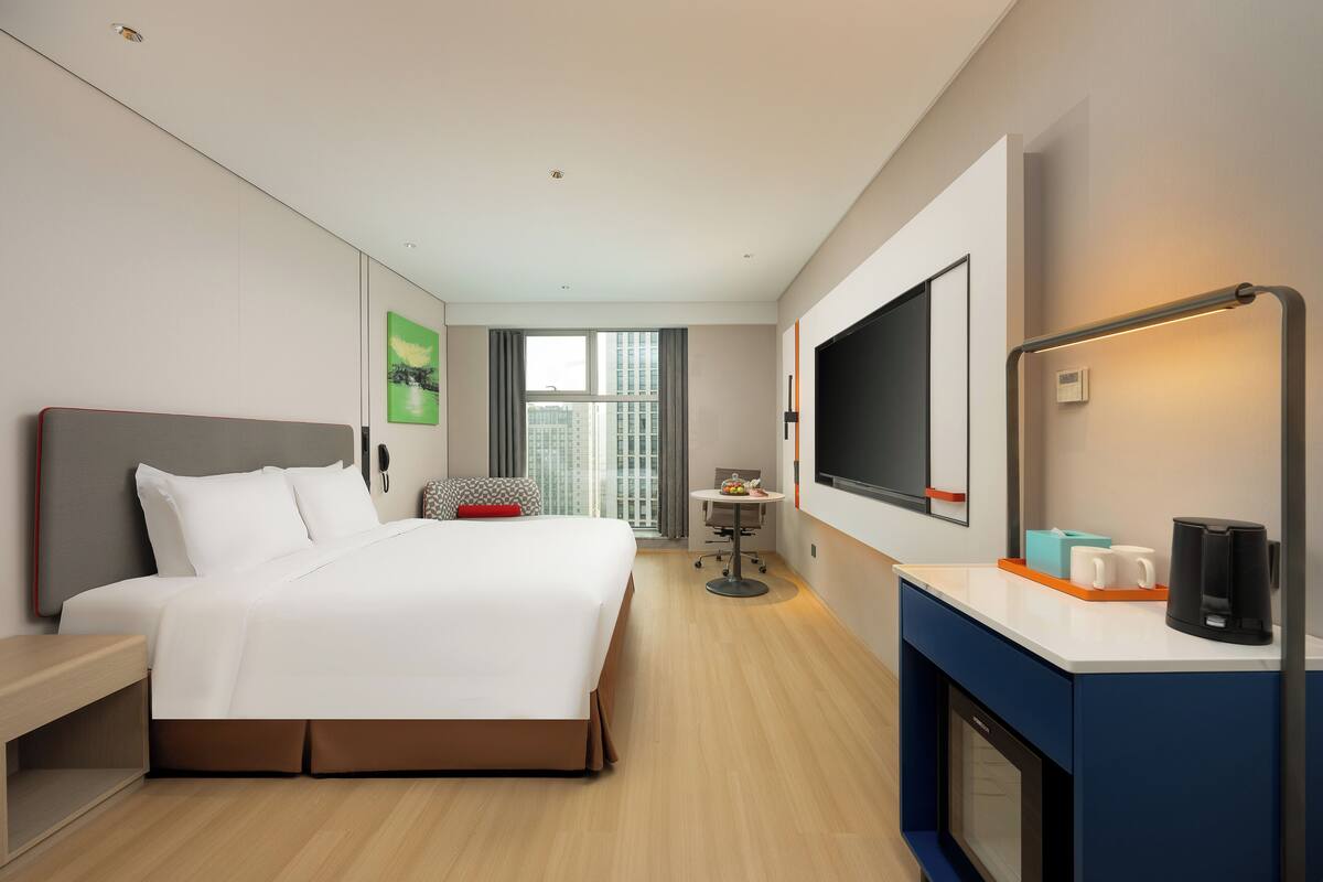 Quarto de hotel moderno com uma cama grande, uma mesa com cafeteira, uma TV e uma janela com vista para a cidade.