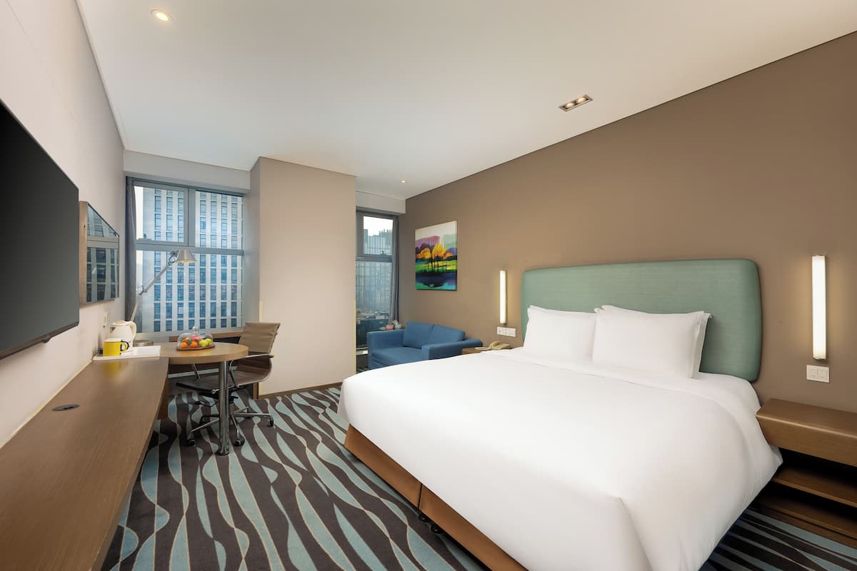 Quarto de hotel com uma cama grande, uma escrivaninha, uma cadeira, uma TV e uma janela com vista para a cidade.