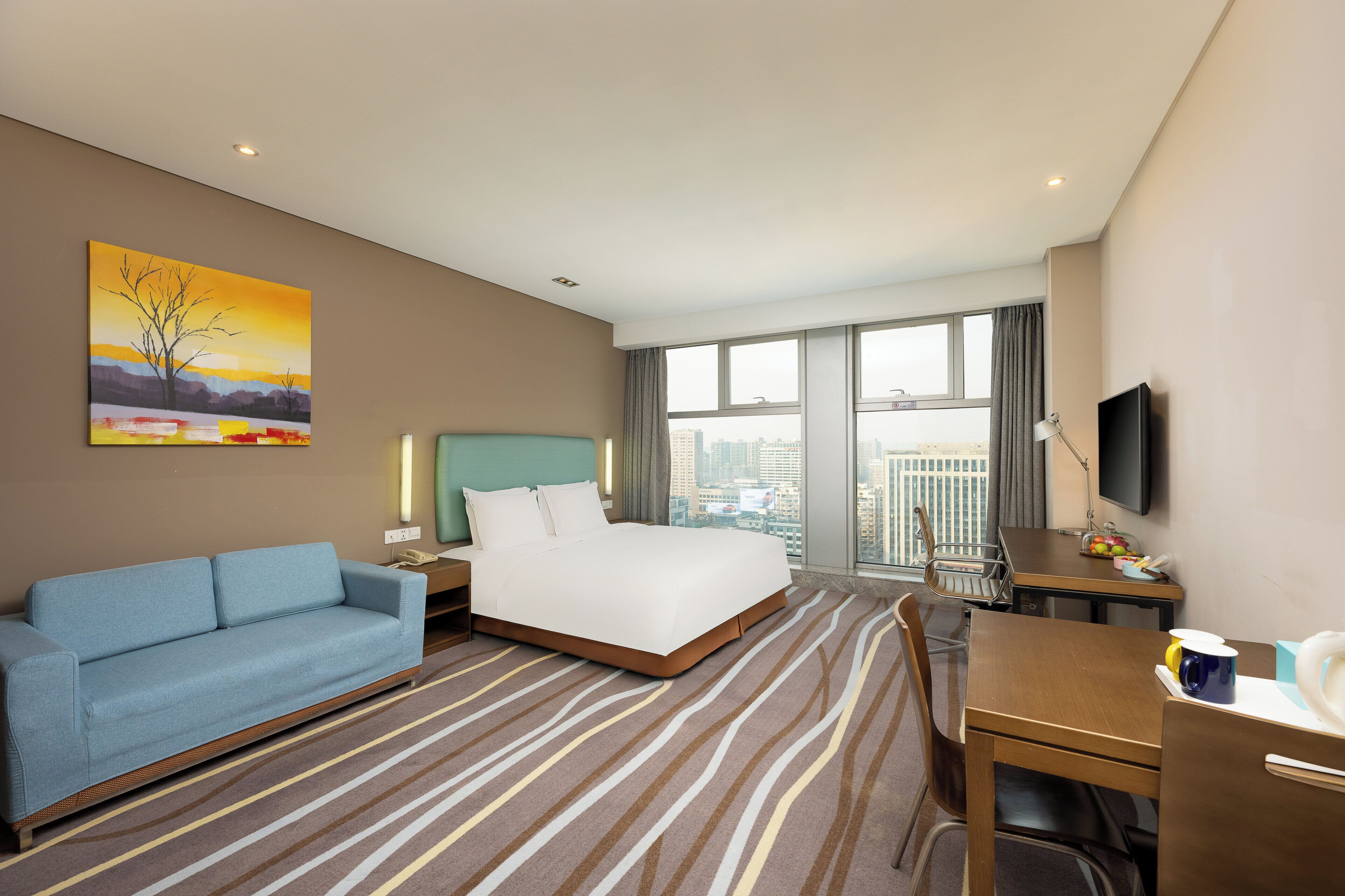 A hotel room with a bed, a sofa, a desk, a television, and a large window with a city view.