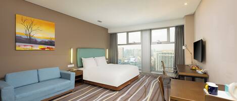 A hotel room with a bed, a sofa, a desk, a television, and a large window with a city view.