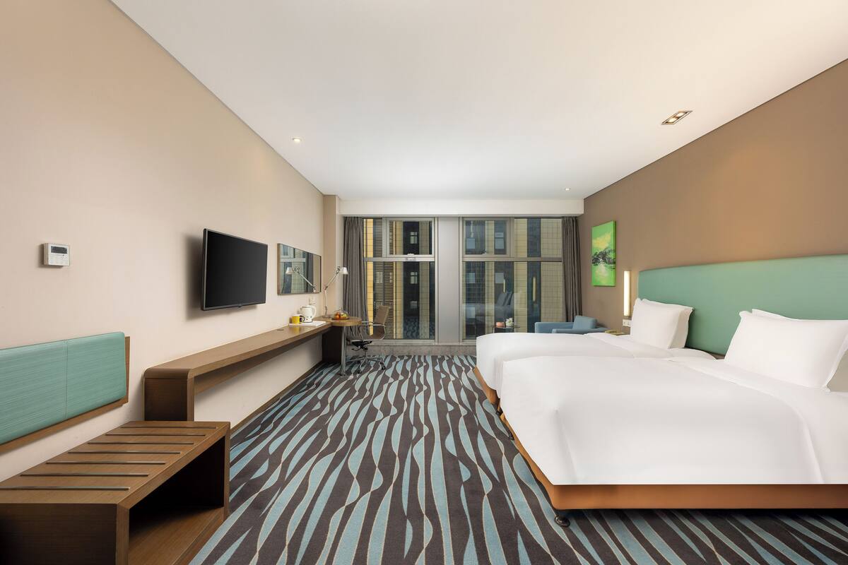 Quarto de hotel com uma cama grande, uma escrivaninha com cadeira, televisão e janela com vista para a cidade.