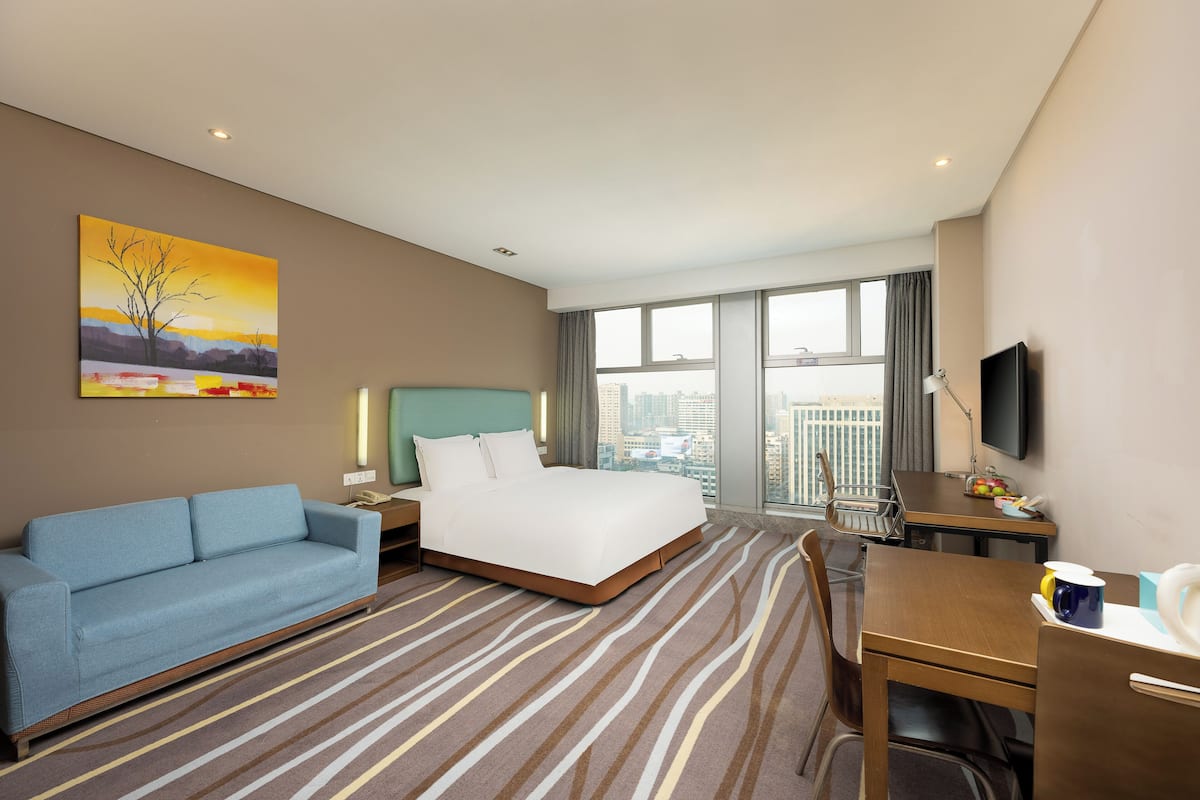 Quarto de hotel com cama, sofá azul, escrivaninha, cadeira, televisão e uma janela grande com vista para a cidade.