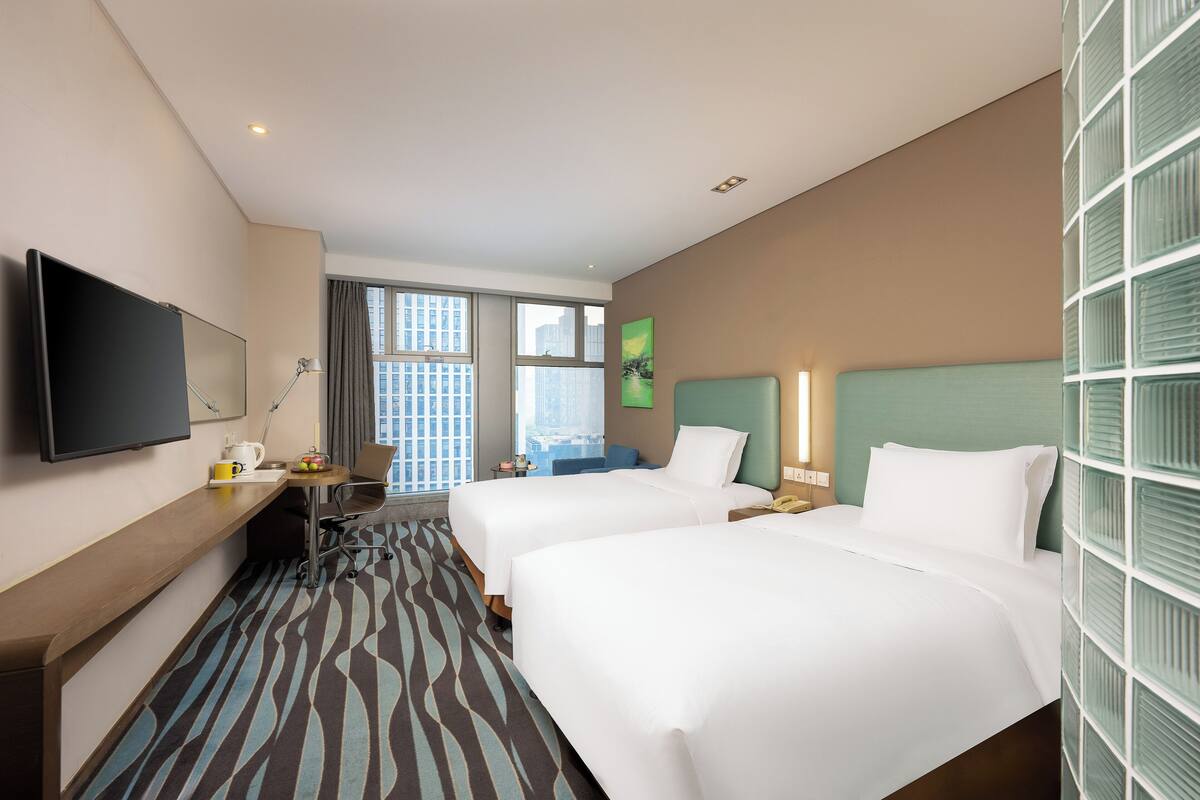 Quarto de hotel com duas camas, uma escrivaninha, televisão de tela plana e janela com vista para a cidade.