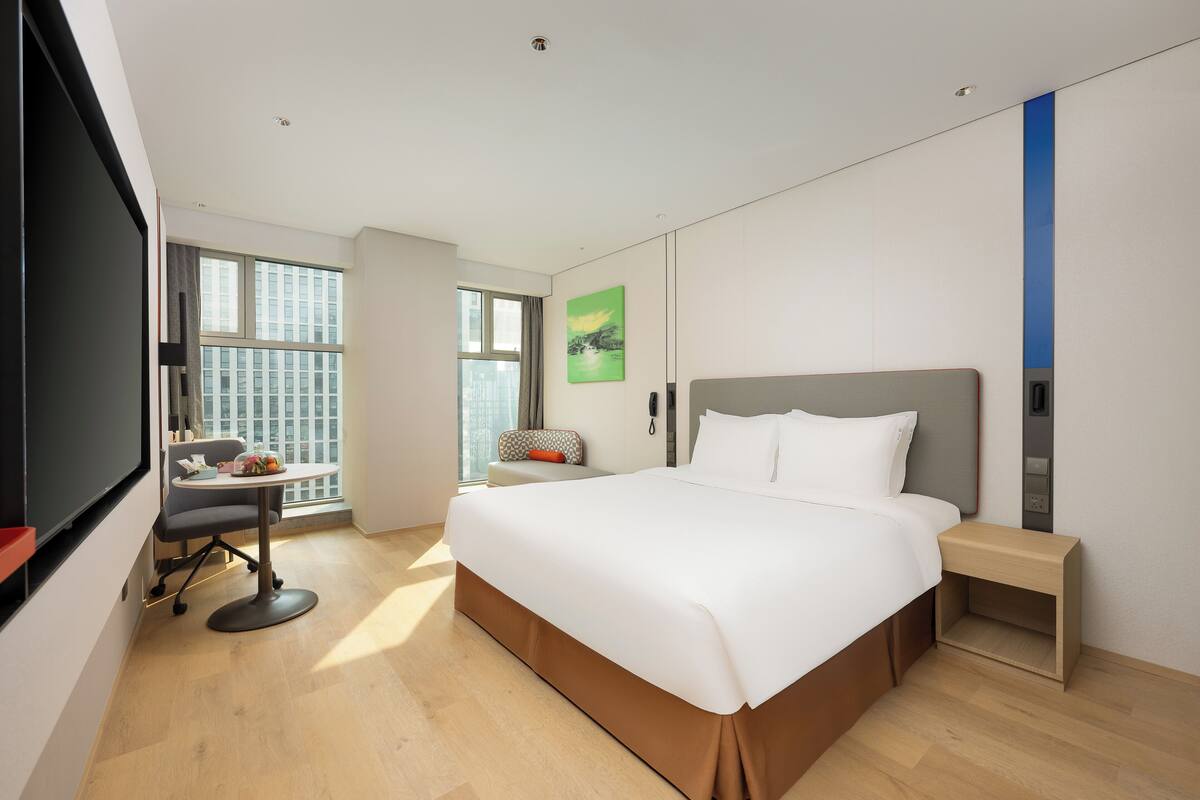 Quarto de hotel moderno com uma cama grande, uma escrivaninha com cadeira, uma mesinha e uma janela com vista para a cidade.