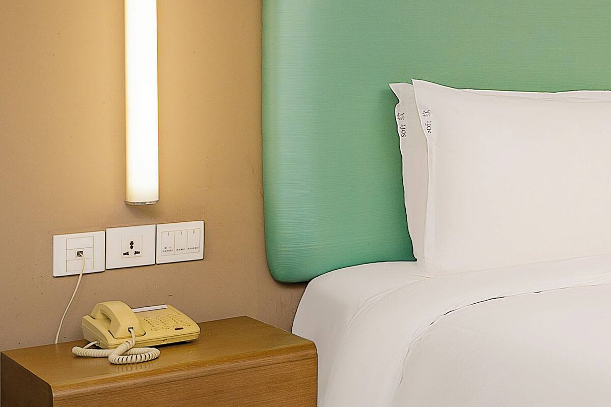 Quarto de hotel com mesa de cabeceira de madeira, telefone amarelo, parede verde e roupa de cama branca.