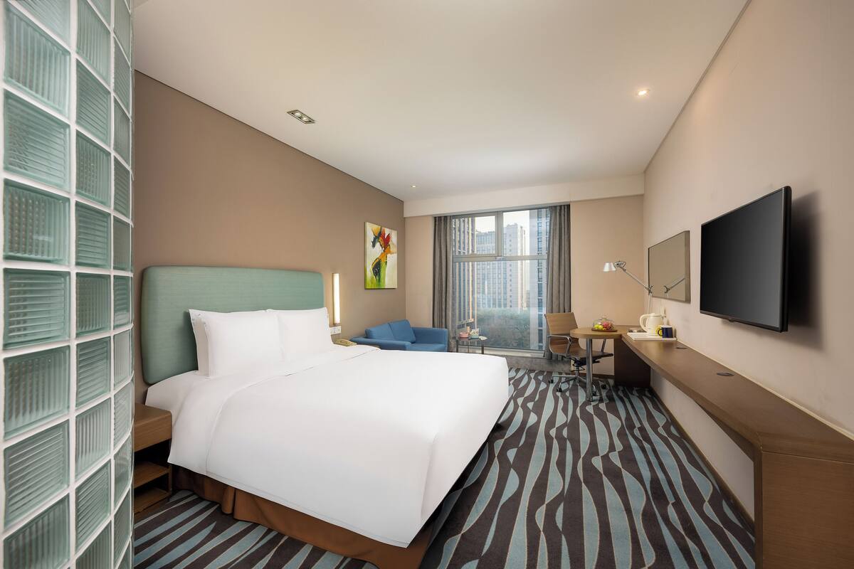 Quarto de hotel com uma cama grande, uma escrivaninha com cadeira, televisão e janela com vista para a cidade.