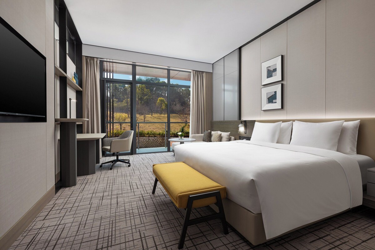 A modern hotel room with a large bed, a desk, a chair, and a view of the outdoors.