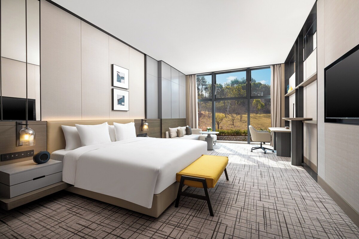 A modern hotel room with a large bed, bedside tables, a desk, and a view of the outdoors.