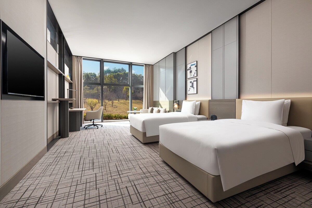 A modern hotel room with two beds, a desk, and a large window offering a view of the outdoors.
