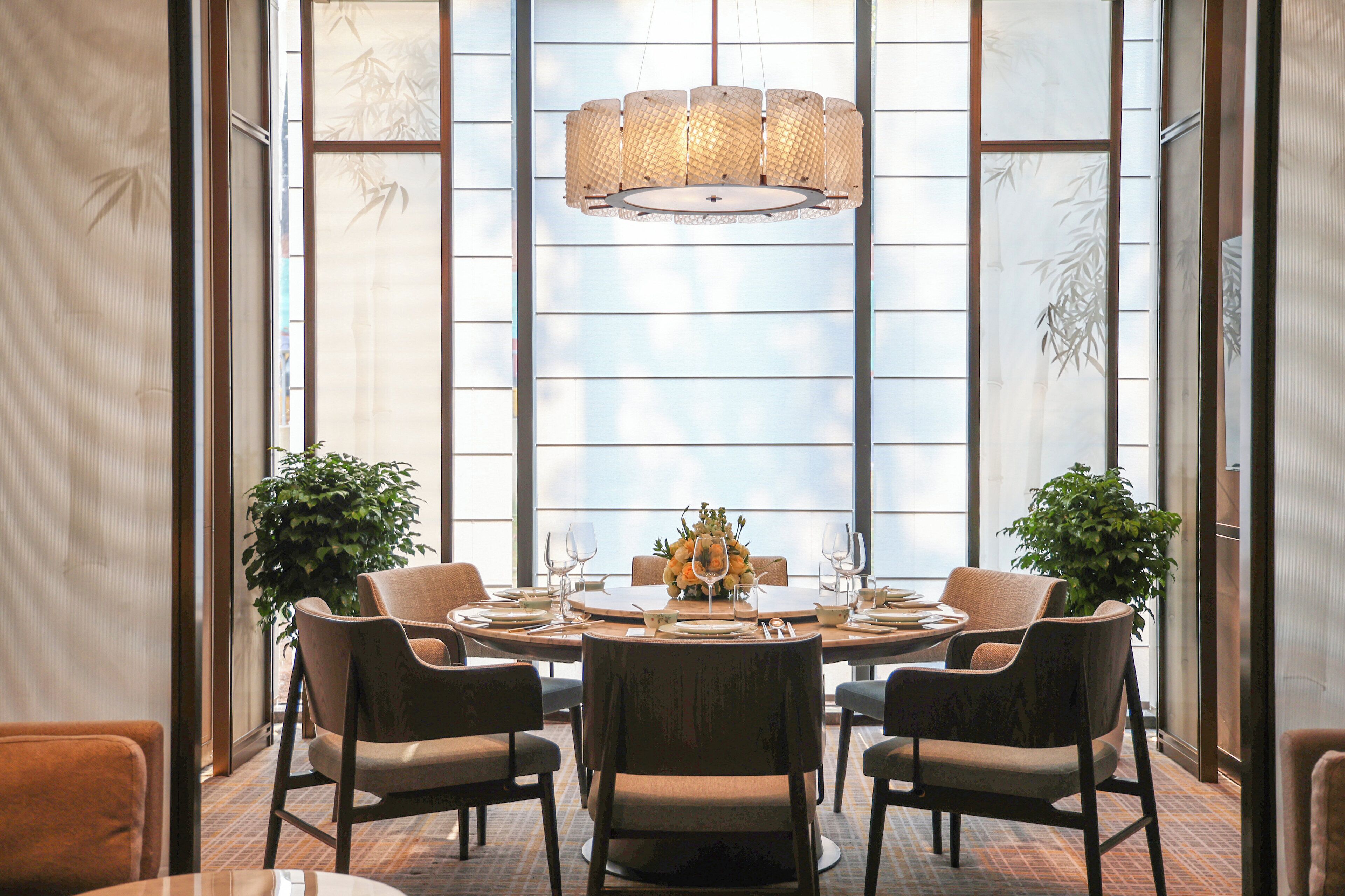 A dining area with a round table set for six, surrounded by six chairs, with a large window in the background and a hanging light fixture above.