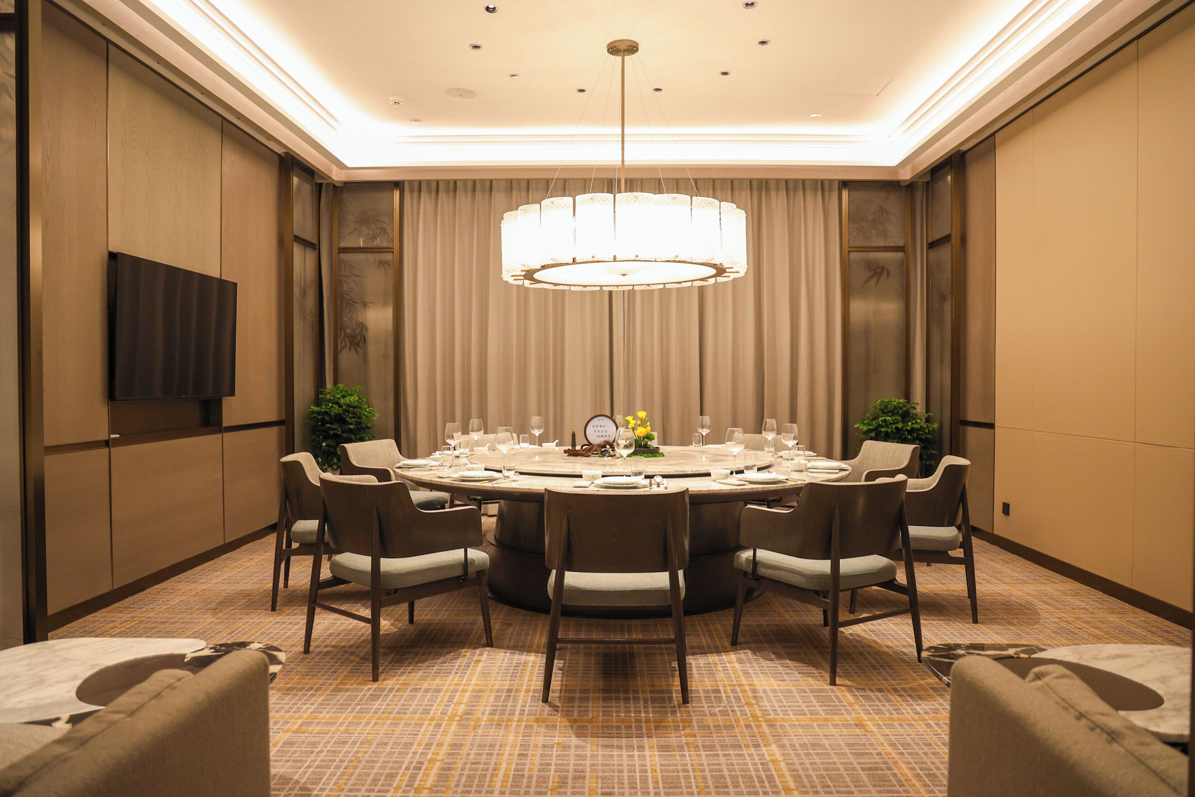 A conference room with a round table, chairs, a chandelier, and a television on the wall.