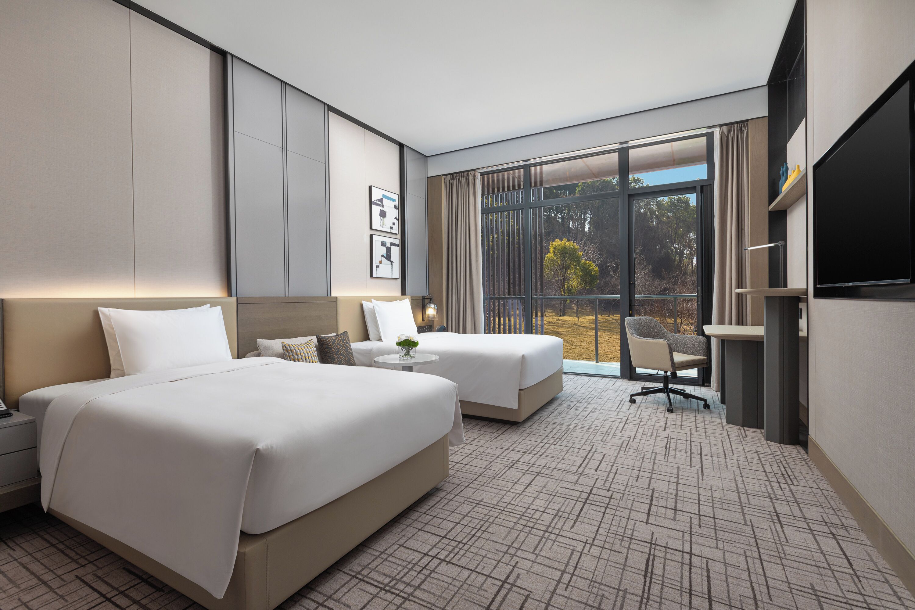 A hotel room with two beds, a desk, and a large window with a view of trees.