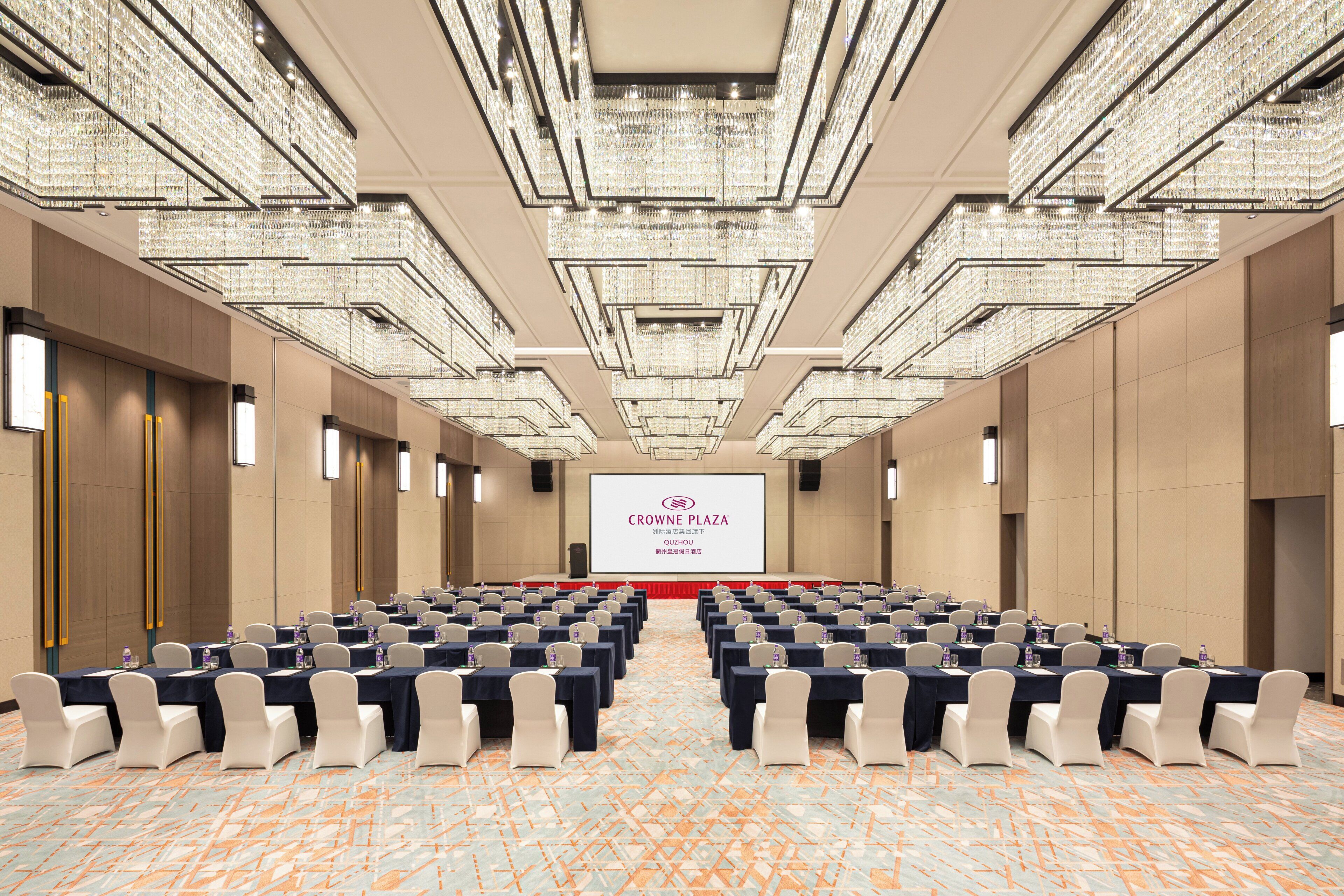 A conference room with rows of tables and chairs, a large screen at the front, and a chandelier ceiling.