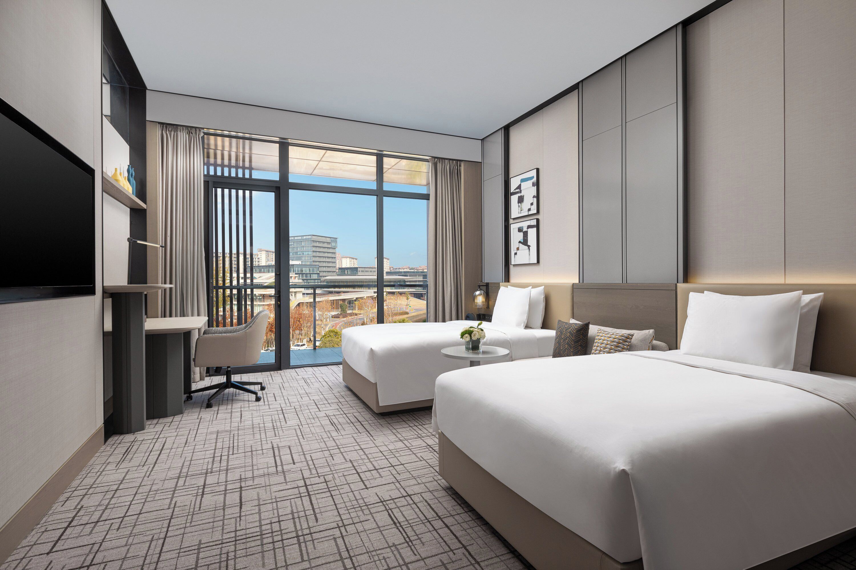 A modern hotel room with two beds, a desk, and a large window offering a city view.
