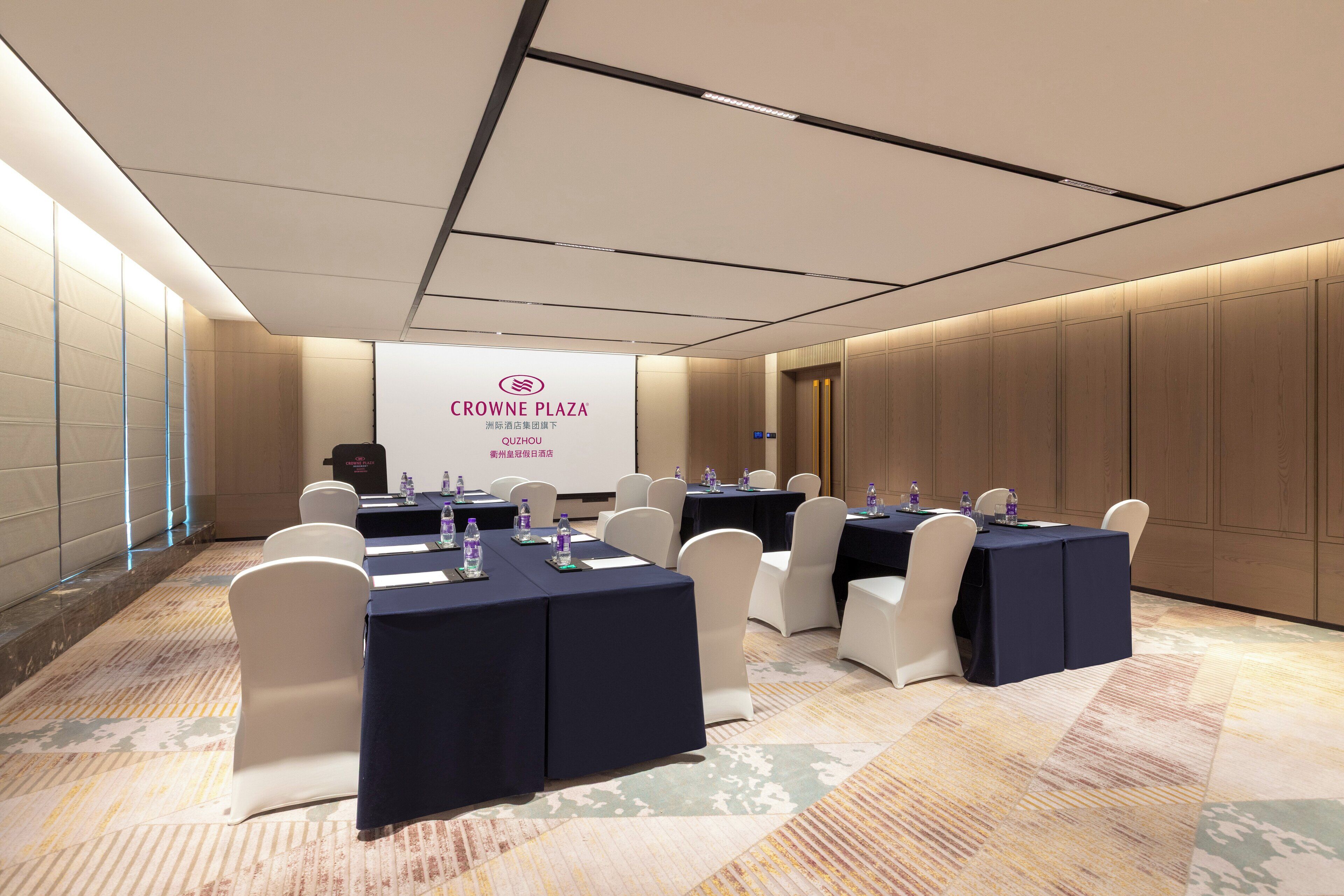 A conference room with tables and chairs, a projection screen, and a sign reading "CROWNE PLAZA".