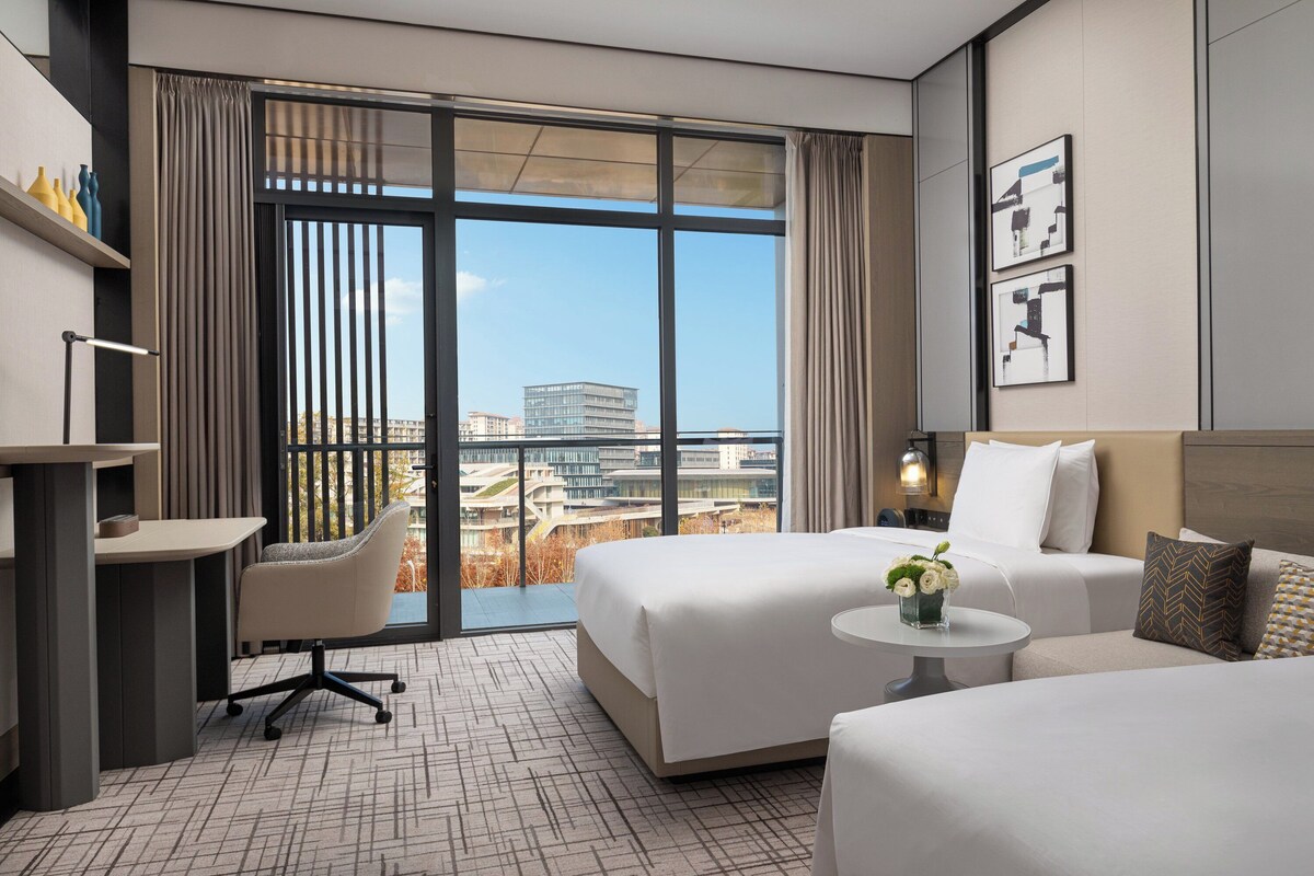 A modern hotel room with a large window, a desk, a chair, a bed, and a small table with a vase of flowers.