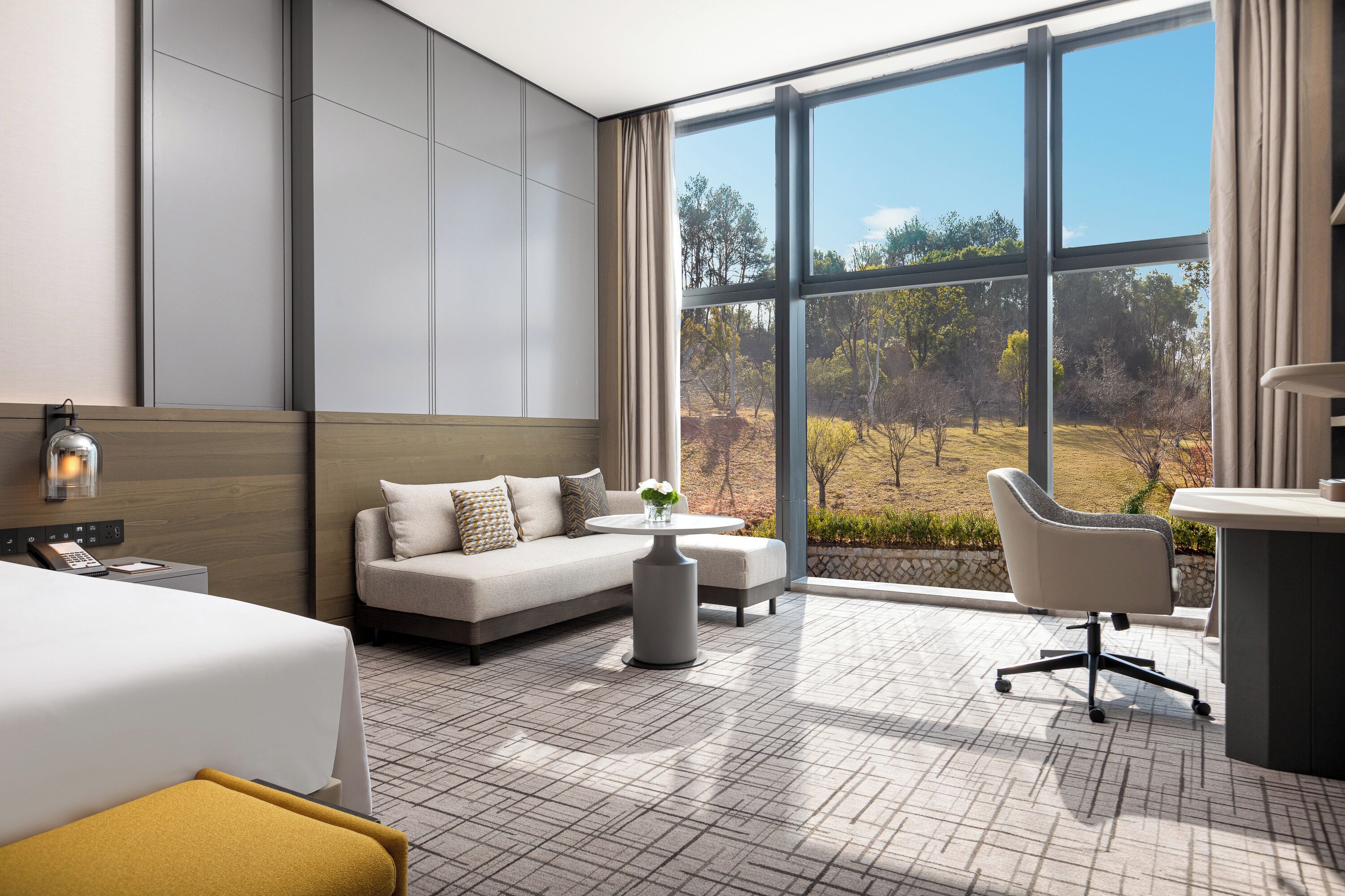 A modern hotel room with a large window, a sofa, a desk, and a bed.