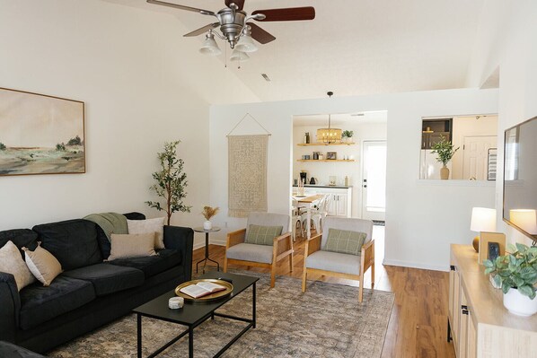 A living room with a black sofa, a coffee table, and a dining area with chairs and a table. There is a ceiling fan, a painting on the wall, and a kitchen in the background. - "The Dawning House" - Peaceful Modern Stay w/ Yard (Georgetown)