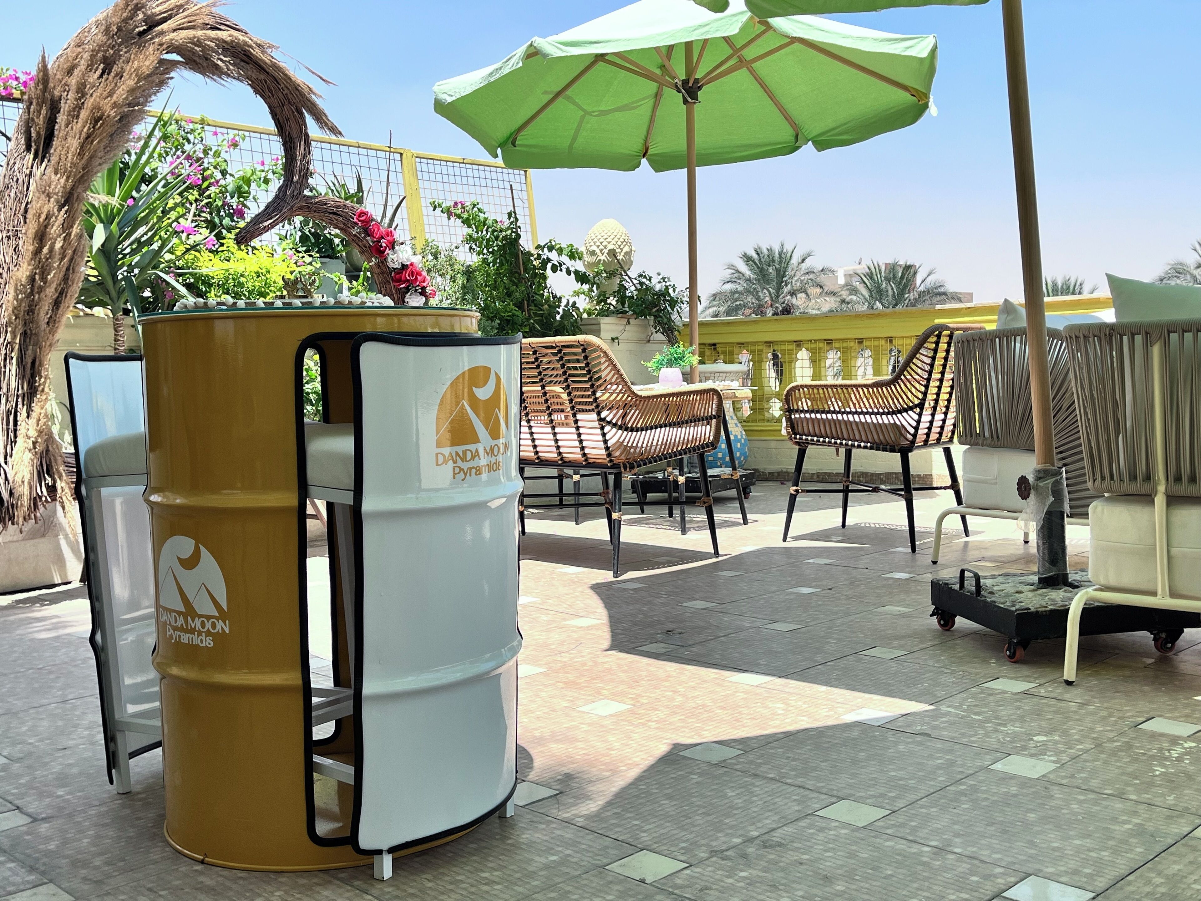 Espace extérieur aménagé avec des chaises en osier et un parasol vert, une poubelle à l’effigie de la marque, et une terrasse carrelée.