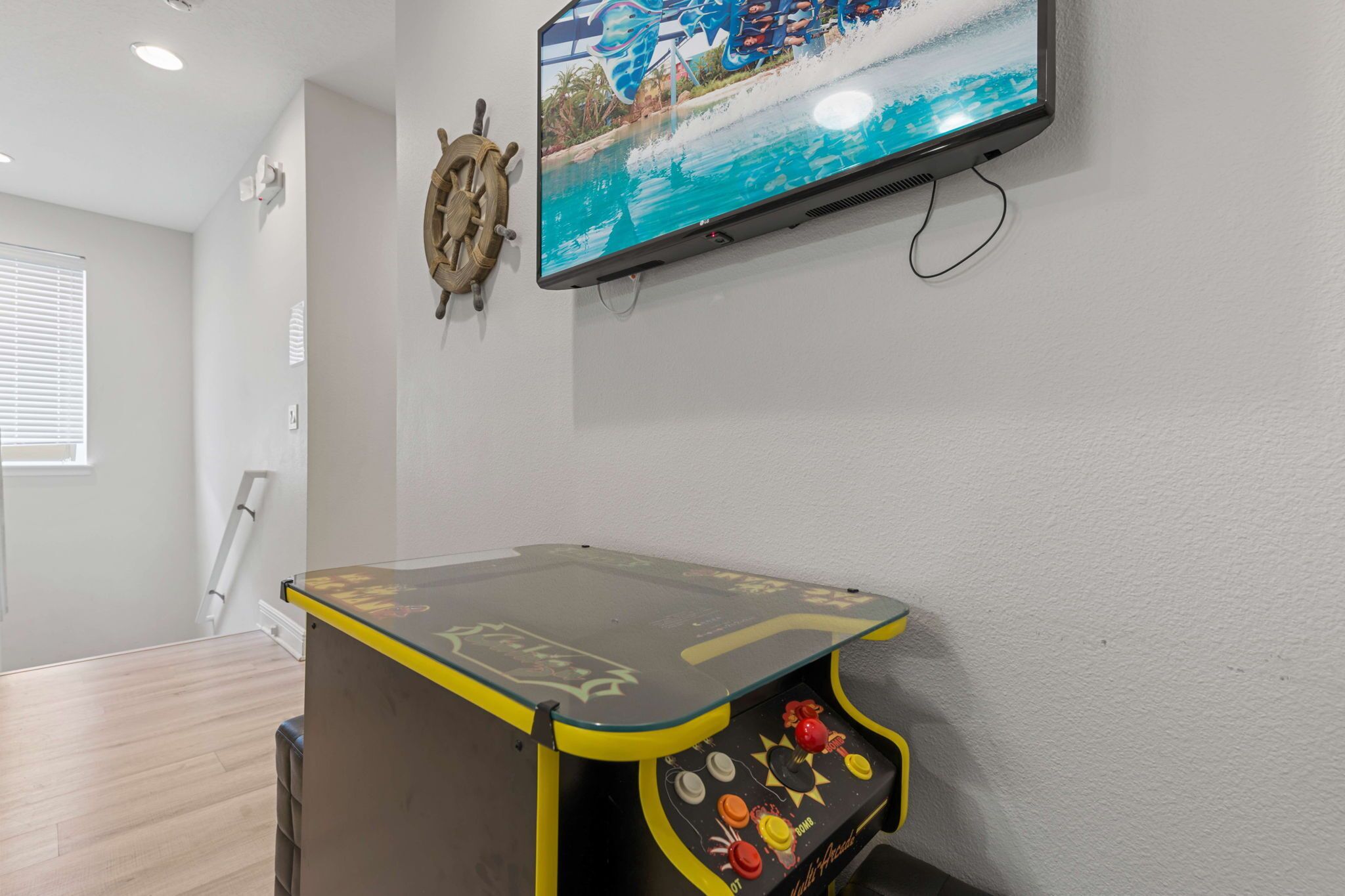 Et værelse med trægulv, et vægmonteret TV der viser et strandbillede, en dekorativt rat og et lille bord med et farverigt kontrolpanel.