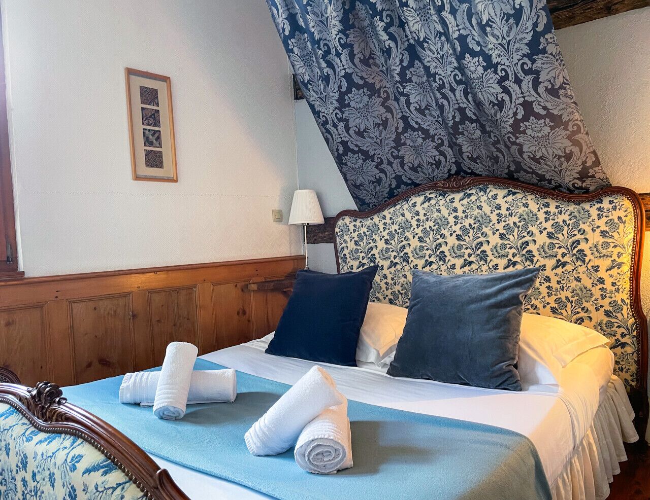 Een net opgemaakt bed met een blauwe spreideken, twee kussens en opgerolde handdoeken.