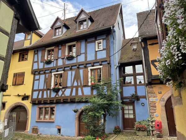 A traditional half-timbered house with a steeply pitched roof, dormer windows, and a small garden area with potted plants and flowers.