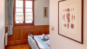 Una habitación con pared de paneles de madera, una cama con sábanas blancas, una ilustración botánica enmarcada y una ventana con cortinas estampadas.