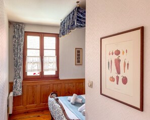 A room with a wooden paneled wall, a bed with white linens, a framed botanical illustration, and a window with patterned curtains.