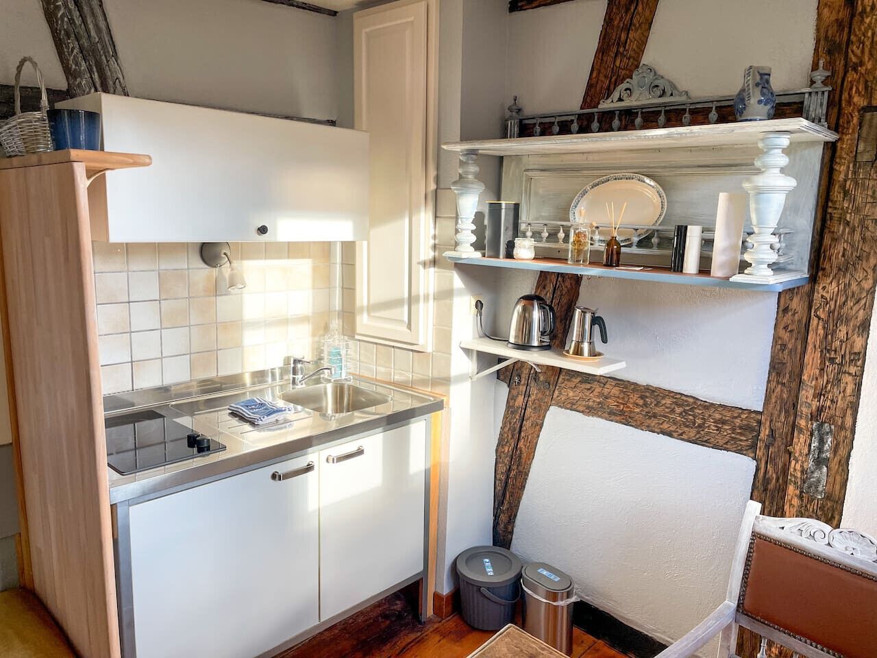 Een compacte keuken met witte kastjes, een roestvrijstalen spoelbak en een houten aanrecht. Er staan twee afvalbakken en een klein tafeltje met een stoel.