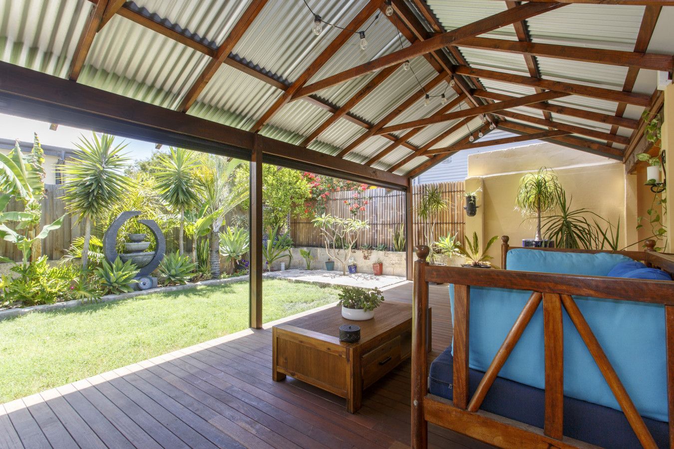 A covered patio with a wooden deck, a seating area with a blue cushion, and a variety of potted plants.