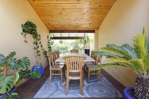A covered patio with a wooden table and chairs, surrounded by potted plants and a patterned rug.