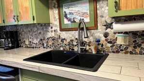 A kitchen with a double sink, green cabinets, a stone mosaic backsplash, and a framed painting above the sink. - Superb 3-bedroom house with WiFi in Neskowin.   (Neskowin)