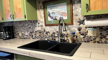 A kitchen with a double sink, green cabinets, a stone mosaic backsplash, and a framed painting above the sink.