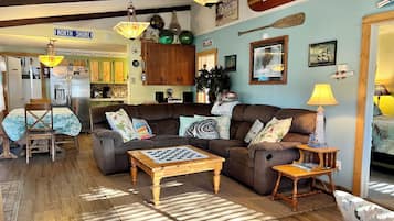A cozy living room with a sofa, coffee table, and a dining area with chairs. There is a kitchen in the background with a refrigerator and cabinets.