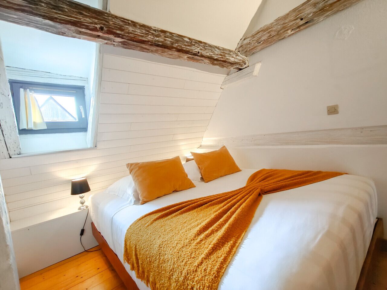Une chambre avec un plafond en bois, une fenêtre, un lit avec une literie blanche et deux oreillers orange, et une lampe de chevet.