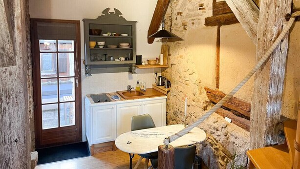 Una cocina rústica con techo de madera, una repisa empotrada con vajilla, una mesa con sillas y una pared de piedra.